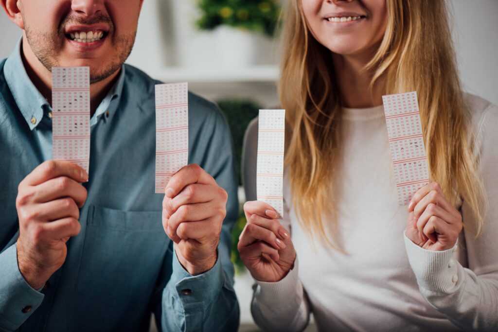 Ein Mann und eine Frau halten mehrere Lottoscheine in die Kamera. Beide lächeln, während sie die Lottoscheine präsentieren.