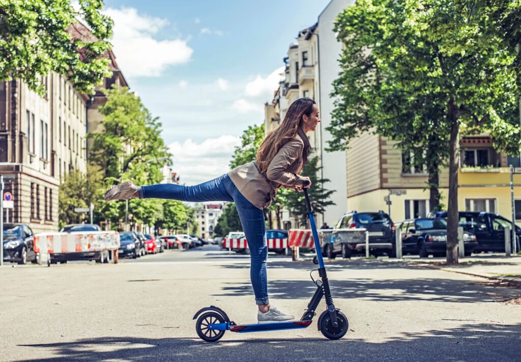 Frau fährt lächelnd auf einem E-Scooter durch eine städtische Straße, während sie spielerisch ein Bein in die Luft streckt.