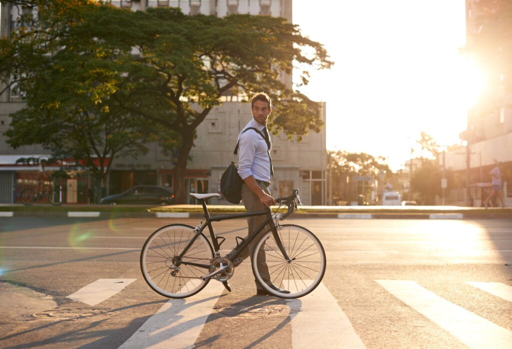 Geschäftsmann hält sein Fahrrad auf einem Zebrastreifen, während er durch die Stadt zur Arbeit fährt, beleuchtet von der aufgehenden Sonne.