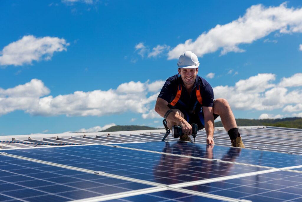 Ein Solarinstallateur montiert Solarmodule auf einem Dach bei strahlendem Sonnenschein. Der Installateur trägt Schutzkleidung und einen Sicherheitshelm. Im Hintergrund ist ein klarer blauer Himmel mit vereinzelten Wolken zu sehen. Die Solarpanels reflektieren das Sonnenlicht und symbolisieren die Nutzung erneuerbarer Energien zur Förderung der Energiewende.