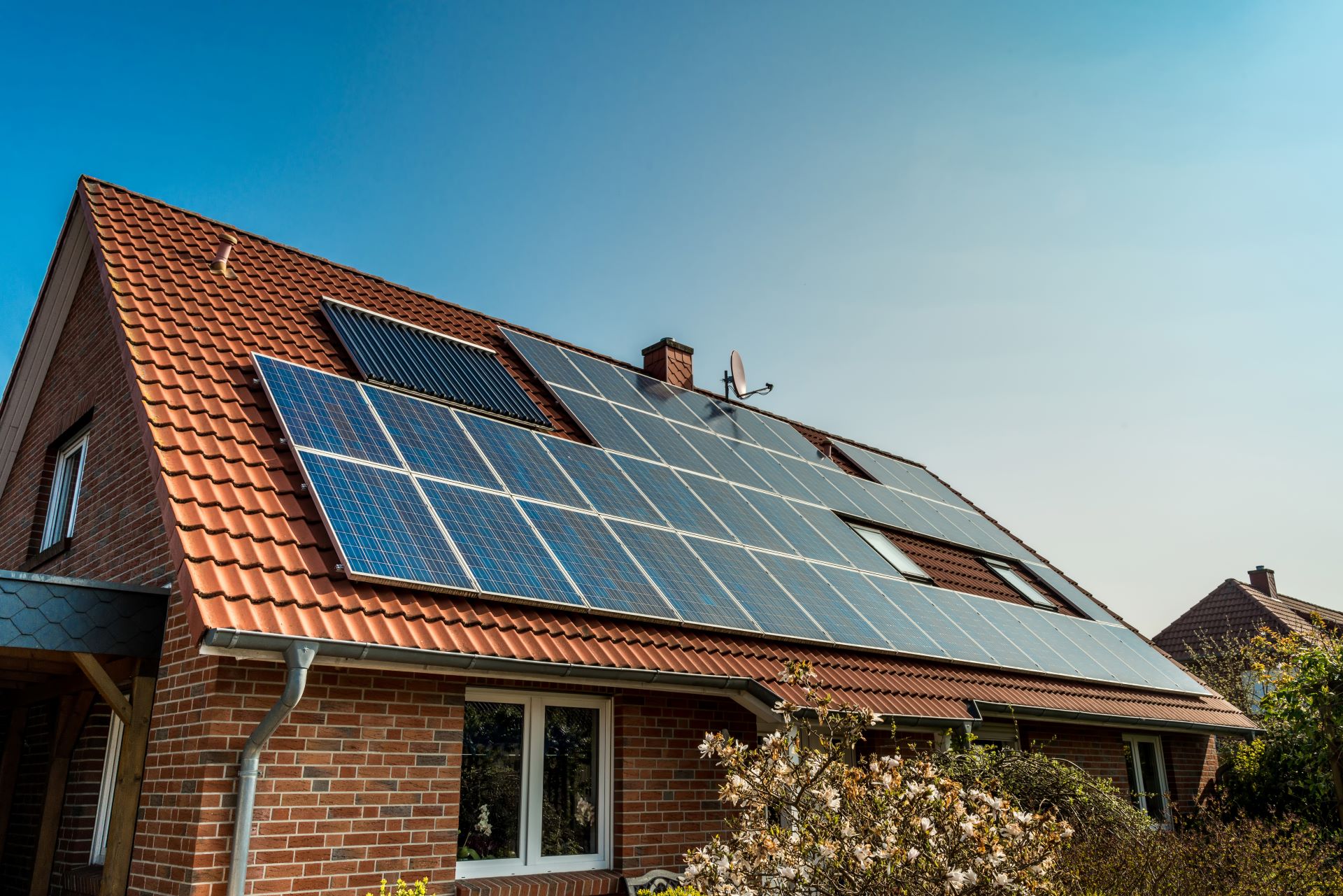 Ein Einfamilienhaus mit einer großen Solaranlage auf dem Dach, bestehend aus mehreren Solarmodulen. Das Gebäude hat ein rotes Ziegeldach, und die Solarpanels sind gleichmäßig darauf montiert. Der klare Himmel und die Sonneneinstrahlung deuten auf eine optimale Nutzung der Sonnenenergie hin. Die Anlage symbolisiert die Integration von Photovoltaik in private Haushalte und den Beitrag zur Energiewende.