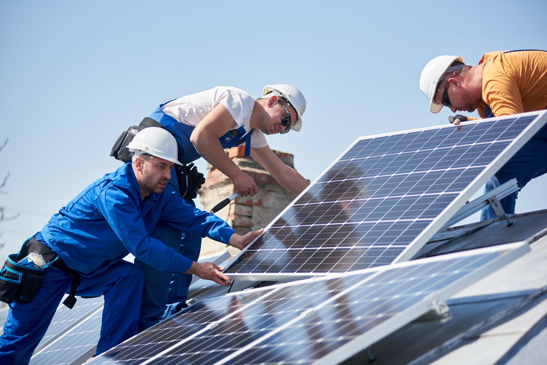 Drei Techniker montieren Solarpanels auf einem Dach. Sie tragen Schutzkleidung und Helme, während sie die Paneele sorgfältig befestigen. Der Himmel ist klar, und die Sonnenstrahlen spiegeln sich auf den Solarpanels. Die Szene symbolisiert den Übergang zu erneuerbaren Energien und die aktive Umsetzung der Energiewende.