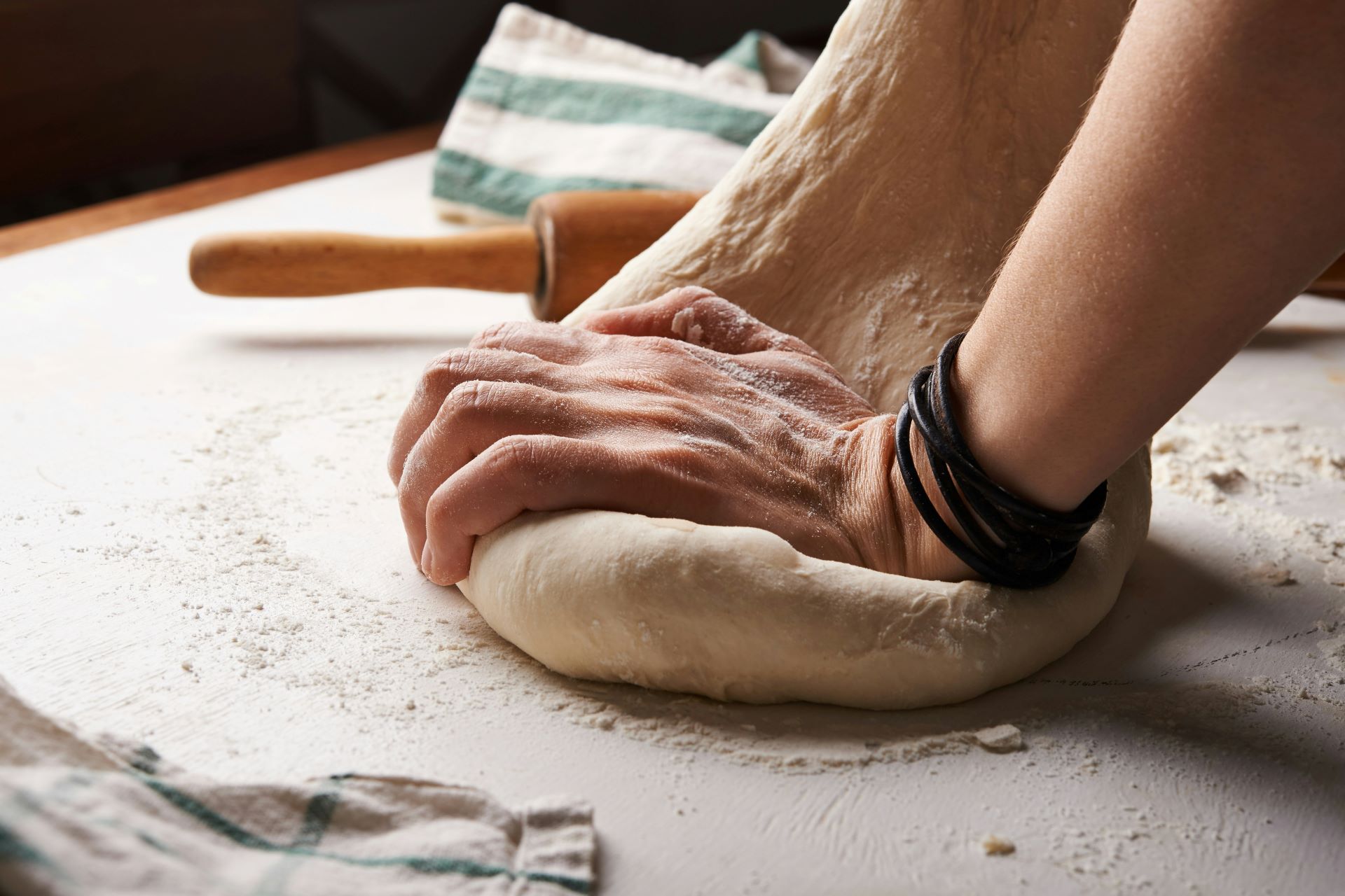 Nahaufnahme von Händen, die einen Pizzateig kneten, mit einem Nudelholz und Küchentüchern im Hintergrund auf einer bemehlten Arbeitsfläche.