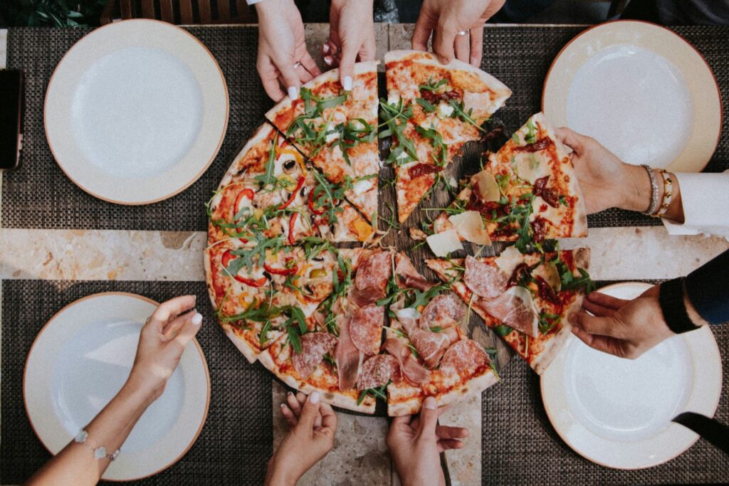 Hände greifen nach Pizza-Stücken auf einem großen, frisch belegten Pizzateller, der auf einem gedeckten Tisch im Freien steht.