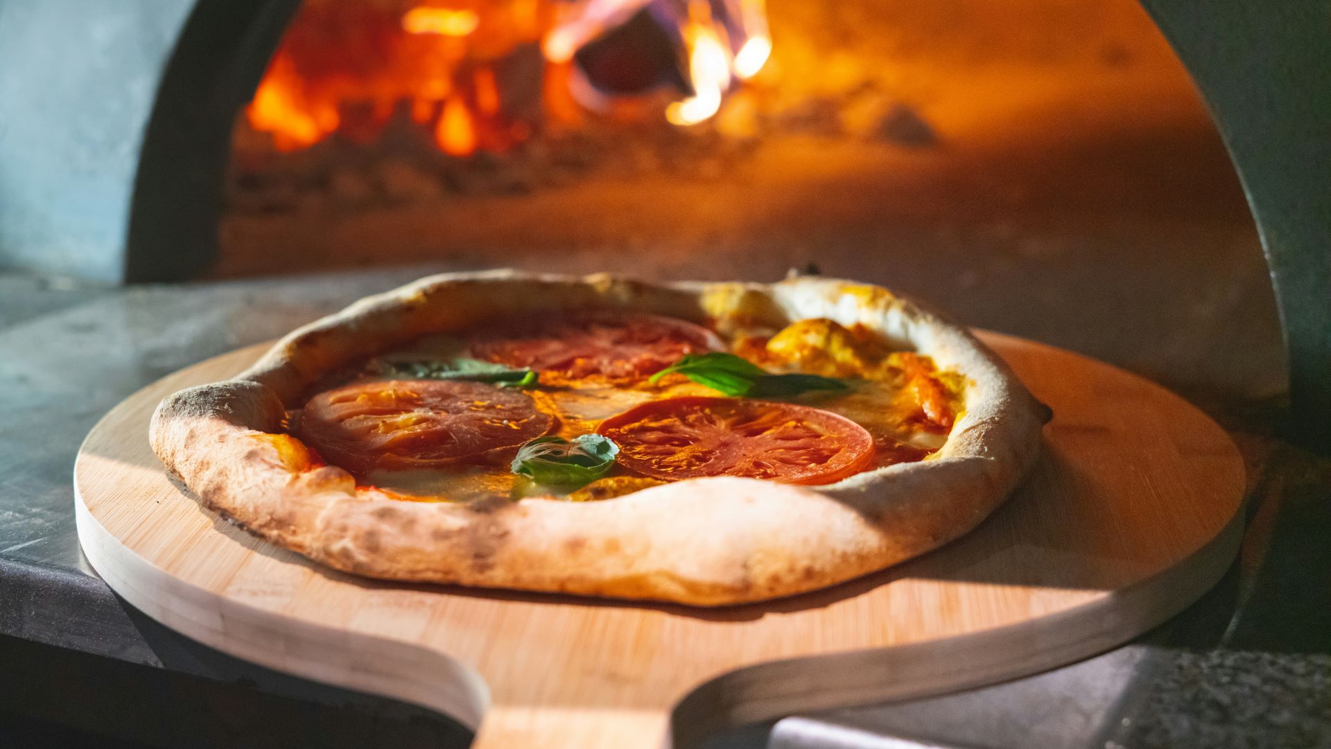 Frisch gebackene Margherita-Pizza mit Tomaten und Basilikum auf einem Holzpizzaschieber vor einem heißen Pizzaofen mit loderndem Feuer im Hintergrund.