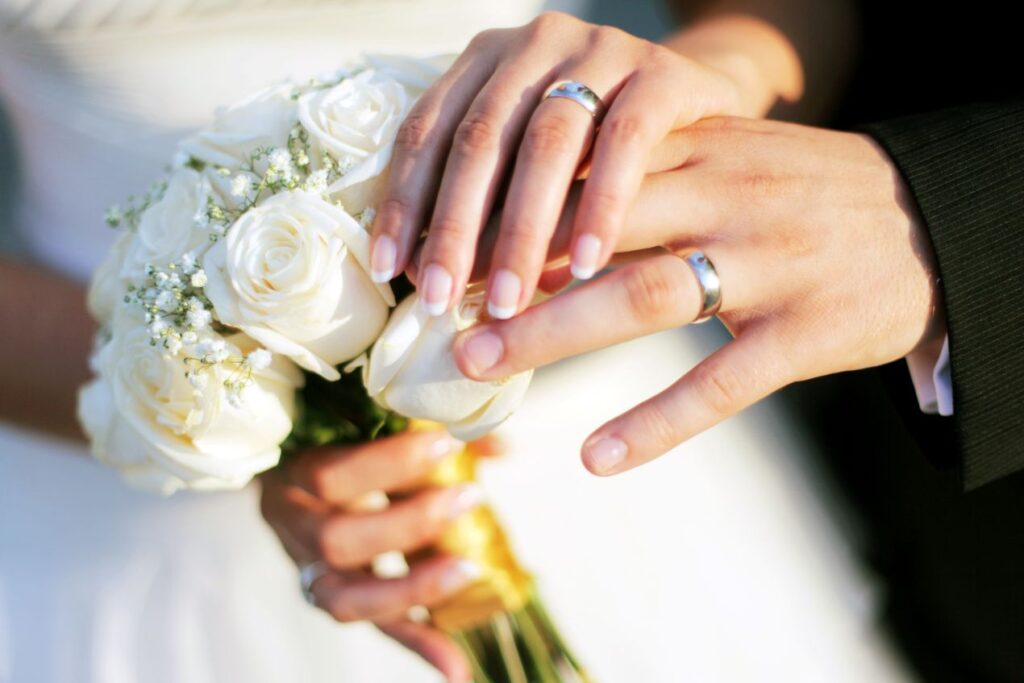 Hochzeitspaar haelt Haende mit Eheringen und weissem Rosenstrauss – Trauring Konfigurator Beispiel