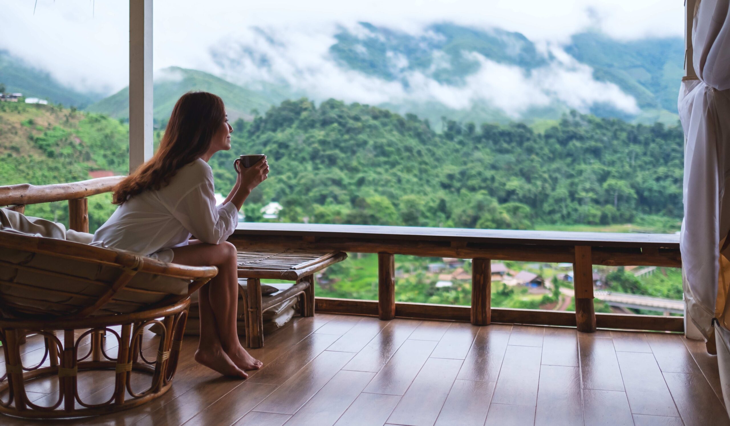 Portraitbild einer schönen asiatischen Frau, die heiße Kaffeetasse hält und trinkt, während sie auf dem Balkon sitzt und auf Berge und die grüne Natur blickt.