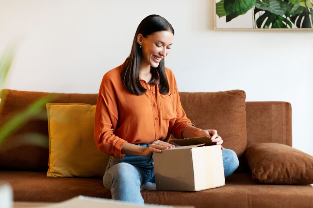 Eine Frau sitzt auf dem Sofa und öffnet ein Paket – Kundengeschenke lösen positive Emotionen aus