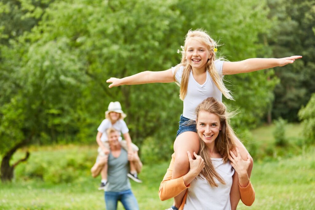 Familie spielt im Gruenen, Kinder auf Schultern der Eltern, glueckliche Momente und Naturerlebnis auf einem Erlebnisbauernhof NRW